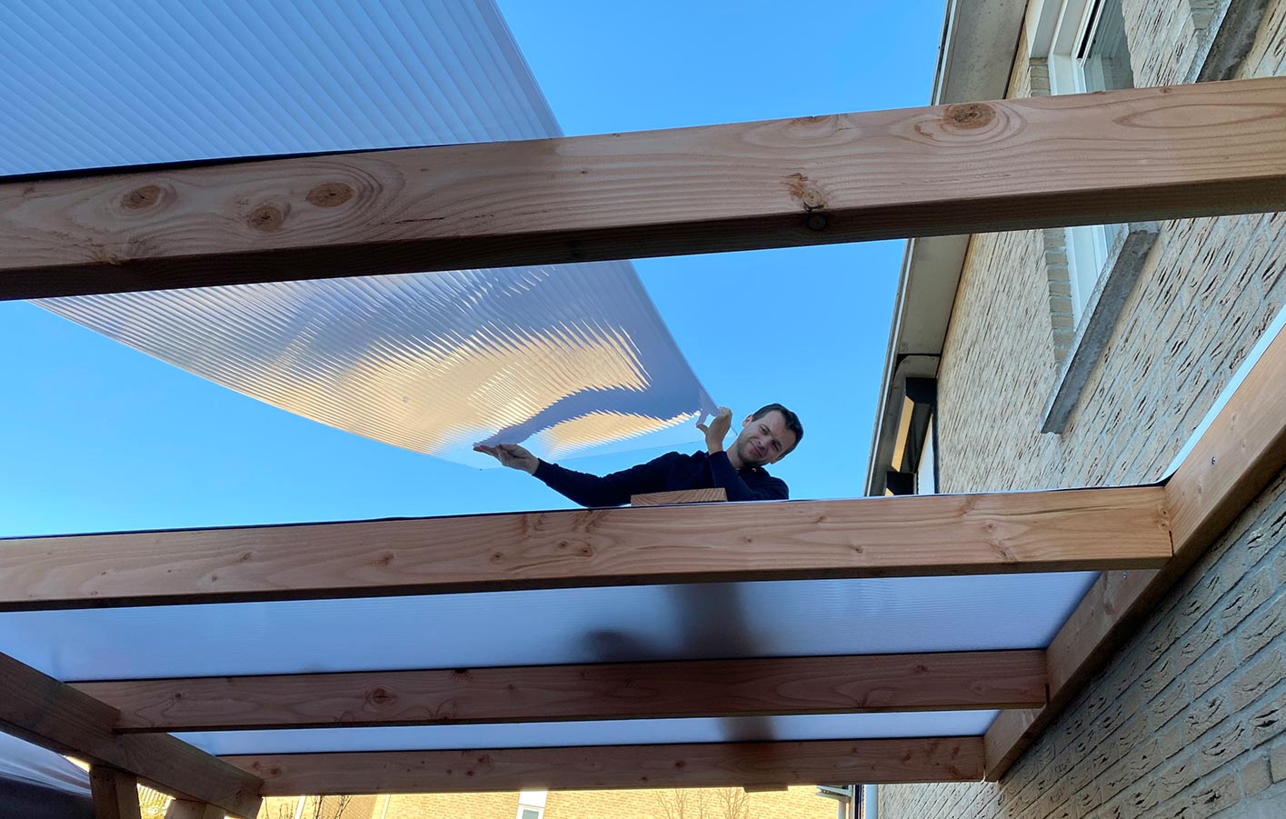 Mann installiert transparente Polycarbonatplatten auf Holzbalken einer Terrassenüberdachung.