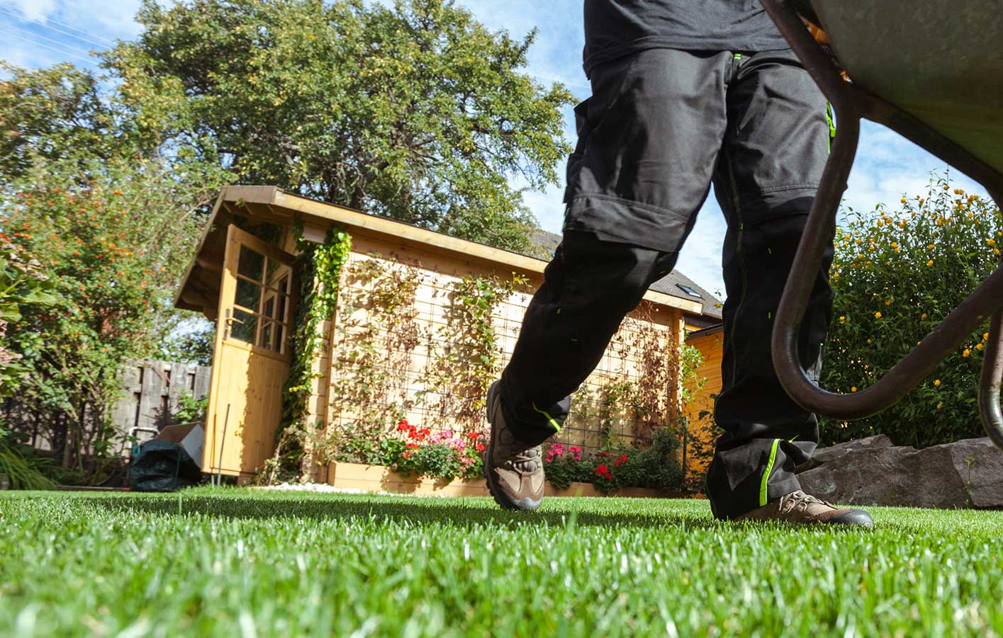Gartenhaus mit Blumenranken im Hintergrund, Mann mit Schubkarre bei der Gartenarbeit im Vordergrund.